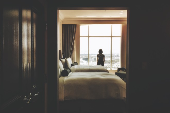 γυναίκα στέκεται όρθια σε δωμάτιο ξενοδοχείου μπροστά από το κρεβάτι κοιτώντας από το παράθυρο
